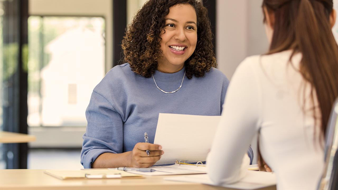 two business women sit across from each other in an office and one is holding papers