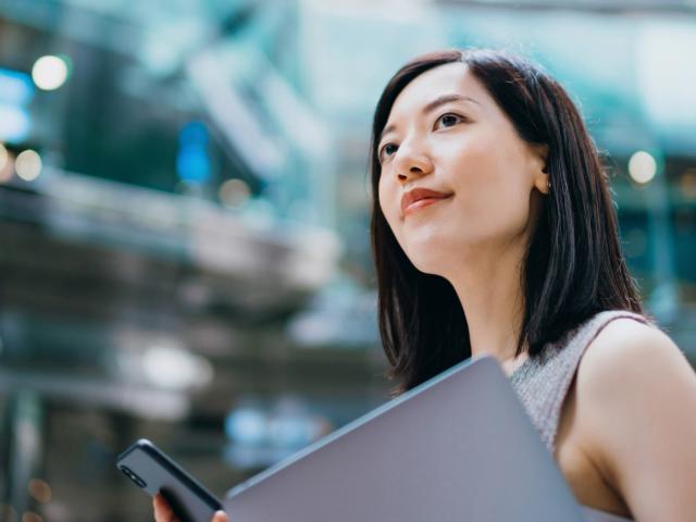 Young professional woman holding laptop, looking up in modern glass office atrium