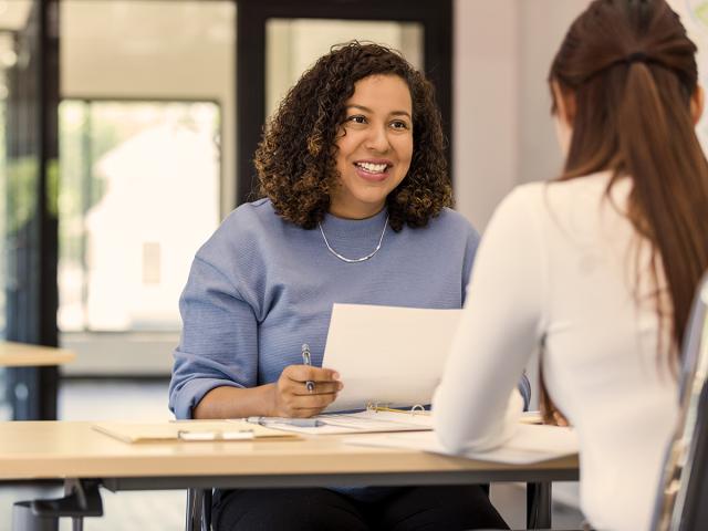 two business women sit across from each other in an office and one is holding papers