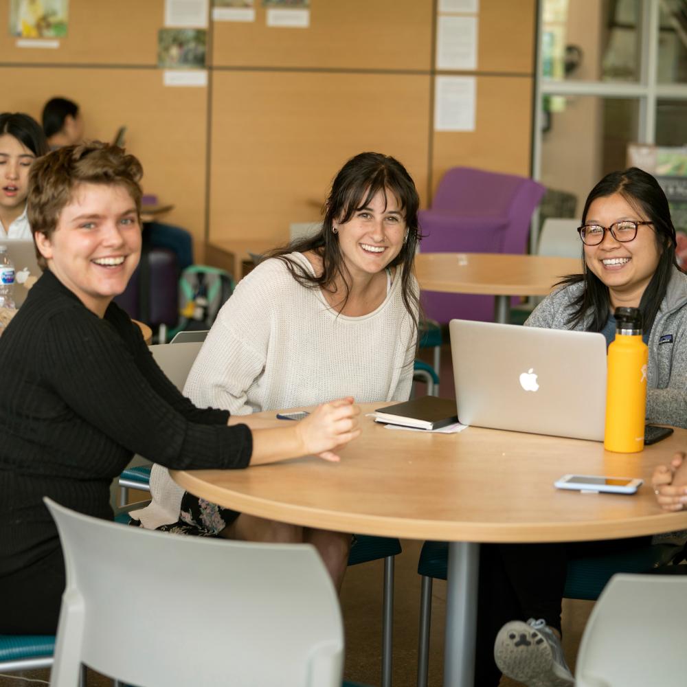 Three women smile at a table with laptops, while others work in a brightly lit room.