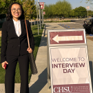 Smiling woman in black suit beside sidewalk sign reading 'Welcome to Interview Day' and arrow