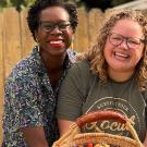 Two women smiling together, holding a basket of colorful fruits in a sunny outdoor setting.
