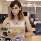 UC Davis undergraduate sits at a table in a bright, modern study space, focused on her laptop. She has light skin, straight brown hair with blonde tips, and wears a white tank top and a lightning bolt necklace. Her laptop is covered with colorful stickers, including animals, food, and slogans like “Black Lives Matter.” Other students are visible in the background, working or socializing in small groups. 