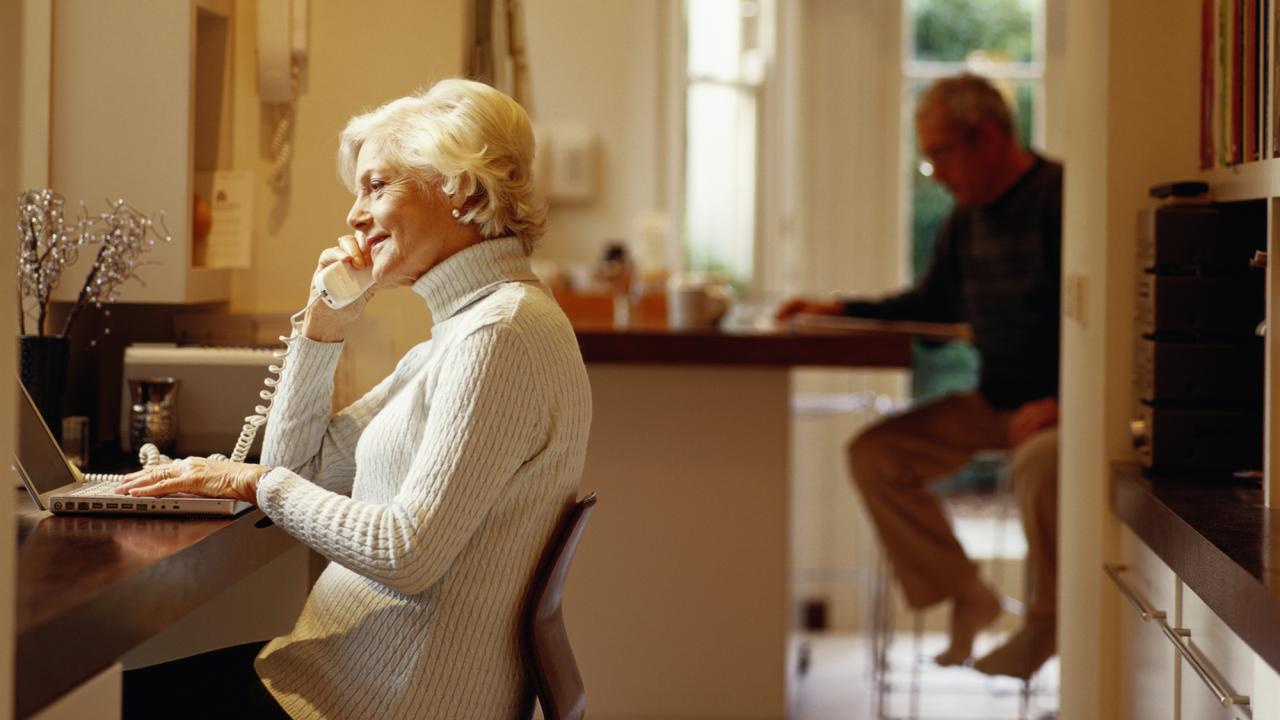 Senior woman using computer and telephone, senior man in background 