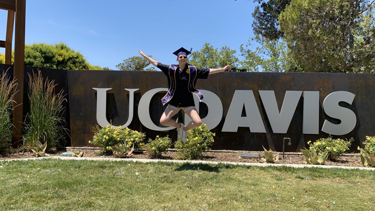 Ava Kemper poses in her graduation cap and gown in front of the UC Davis sign