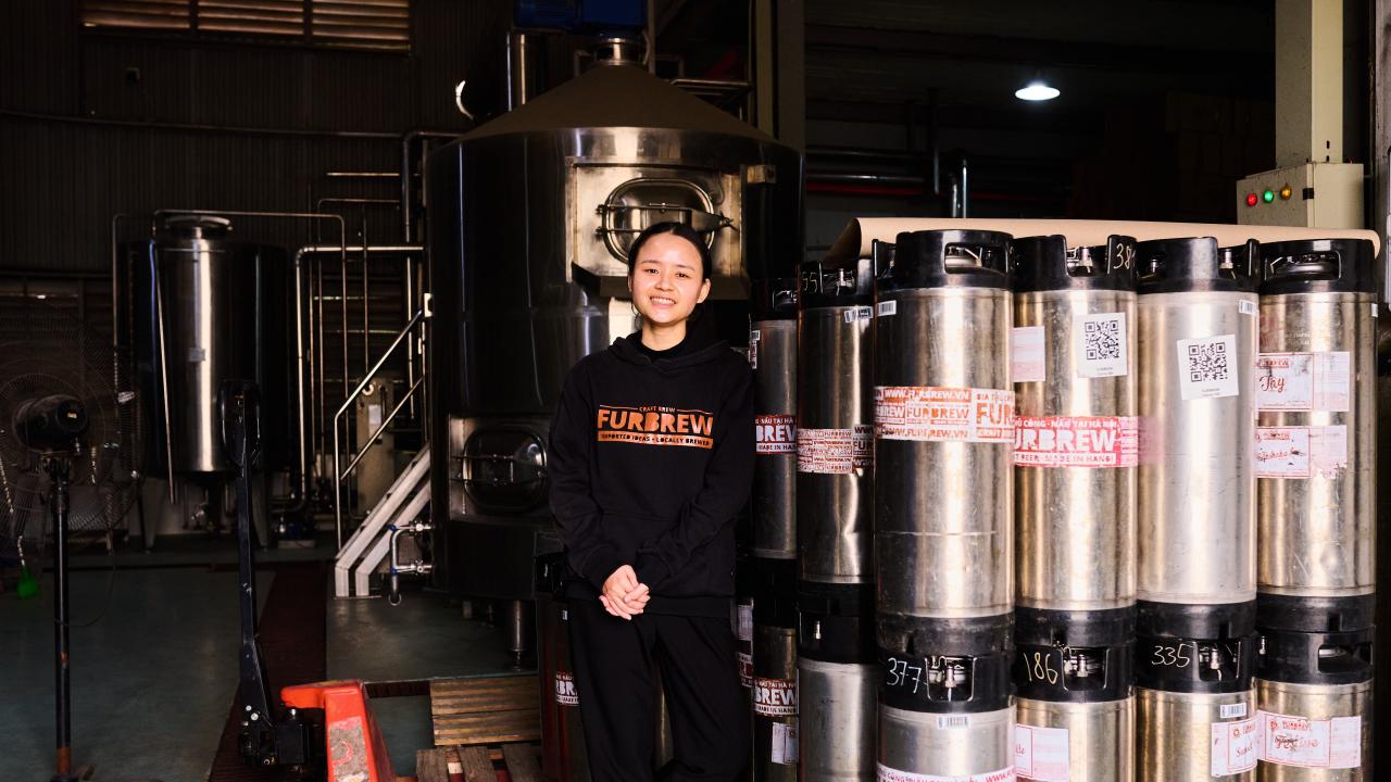 Tapping Potential scholarship winner Dung Ngô stands alongside canisters of beer and other brewing equipment. She wears a black sweatshirt  that reads "Furbrew" in orange lettering.
