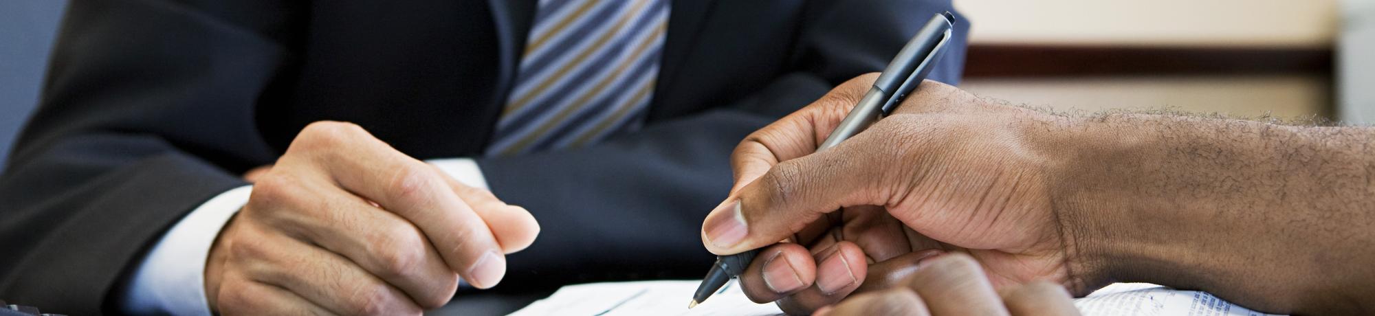 Close-up shot of a pair of hands signing what looks to be an official document., while someone in a suit looks on.